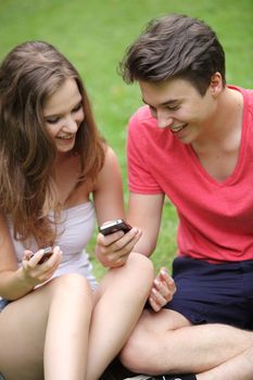 Happy couple of beautiful Caucasian teenagers looking at their mobile phones sitting on grass