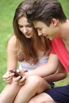 Young students, a teenage boy and girl, sitting on the grass reading a text message on their mobile phone