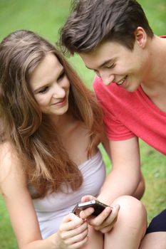 Smiling young teenage boy and girl sitting on green grass using a mobile phone reading text messages