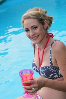 Beautiful woman enjoying a drink at the pool sitting in her bikini smiling at the camera with a glass in her hand