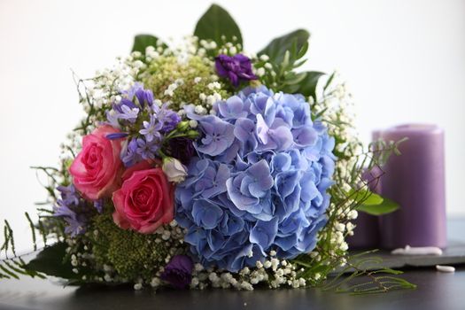 Unusual bridal bouquet with pink roses and blue hydrangeas lying on a table alongside a candle