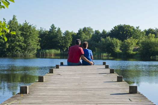 Father and son sitted on a wooden pier on a quiet blue pond in Denmark