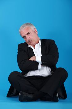 middle-aged man sitting cross-legged and cross-armed against blue background