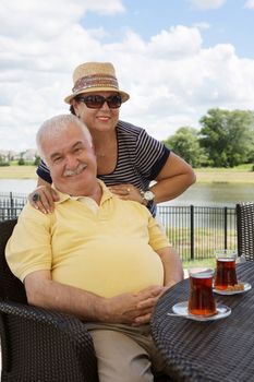Loving senior couple having drinks on an outdoor patio giving the camera beautiful beaming smiles with the woman posing behind her husband with her hands on his shoulders
