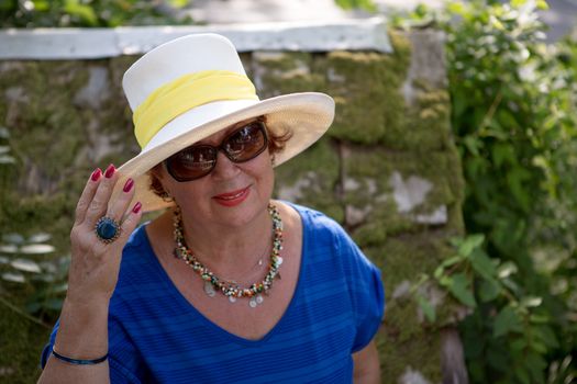 Senior lady with the hat and glasses infront of the greenery posing.