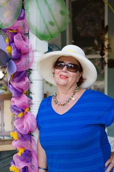 Stylish senior woman in sunglasses and a wide brimmed straw sunhat leaning against a pole alongside decorative pink flowers