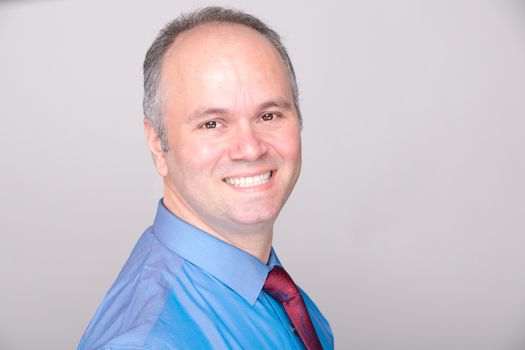 A middle age office worker smiling to the camera with his blue shirt and red tie.