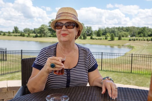 Stylish senior woman in a hat and sunglasses enjoying a drink outdoors sitting at a table overlooking a lake