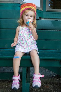 Cute little blond girl in a straw hat sitting on a wooden bench eating a colourful ice cream in the summer sunshine