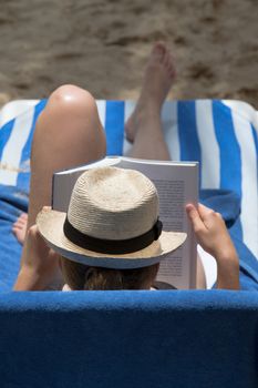 Lady in hat reading her book  on the beach recliner