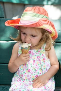 The blonde girl is wearing a floral dress, colorful hat and is eating an ice cream.