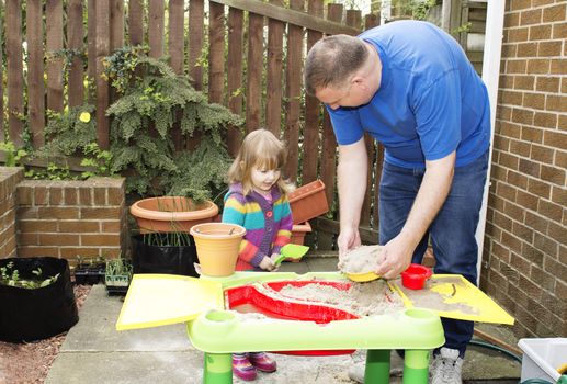 A father and small child are playing together at a sand table in a garden setting
