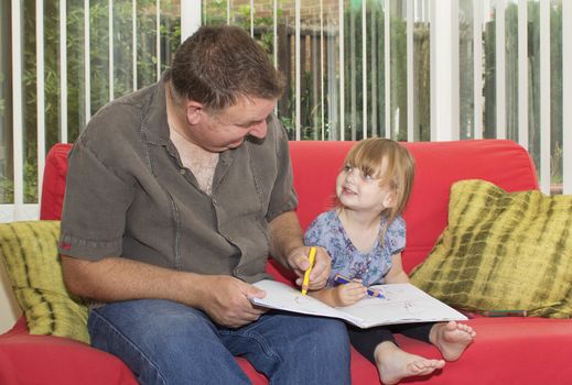 Father and daughter sitting on a sofa, drawing a picture together. The little girl is smiling up at her dad.