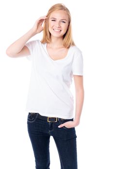 Happy young blond female student in casual jeans standing facing the camera with her hand raised to her hair smiling at the camera, threequarter body on white