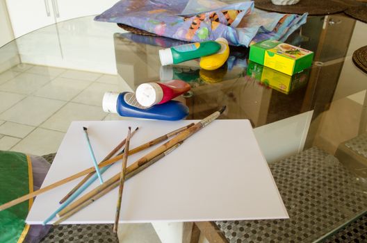 on a glass table to prepare painting supplies white paper of varying size brushes gouache box and liquid spray paint tubes