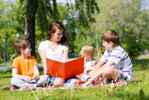 children and teacher reading book together in the summer park