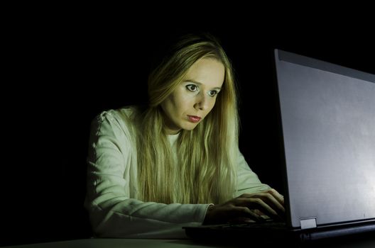 woman working on a computer by night in a dark room with only light from computer falling on her face horizontally cropped