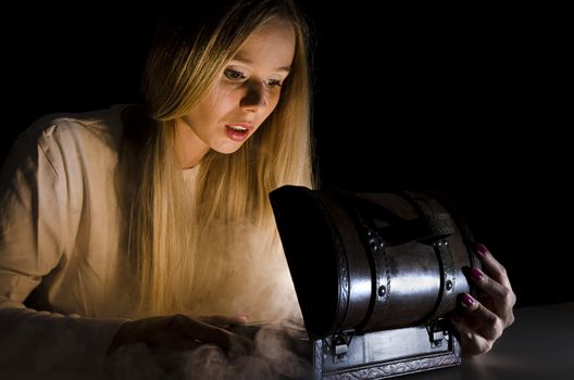 woman secretly opening a small chest in the dark with smoke and lights coming from it and falling on her face