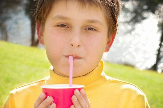 A young boy sips strawberry milkshake outdoors.  He is wearing a yellow skivvy and is backlit by the sunshine.  Some texture added.