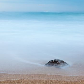 Photo of a rock on a beach, buried in the sand and covered by the ocean. Long exposure. Copy space.