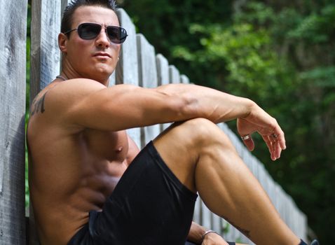 Handsome, serious muscleman sitting against wood fence outdoors, shirtless