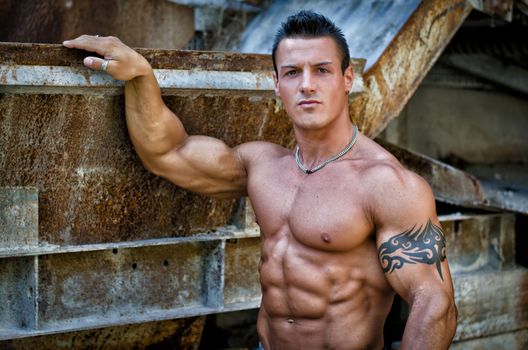 Handsome young muscle man shirtless with hand on rusty metal structure, looking in camera