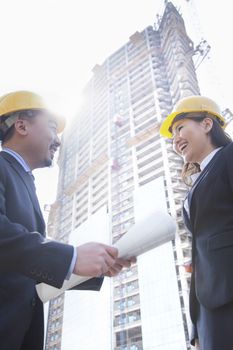 Two architects smiling at a construction site holding blueprint