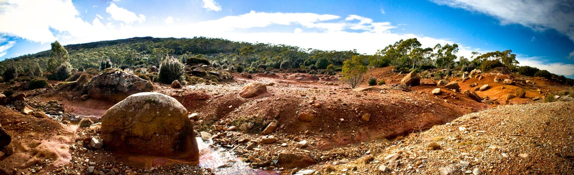 Panoramic scenic view of the Australian outback desert with barren rich red earth and rocks leading to sparse vegetation on the horizon under a hot cloudy blue sky