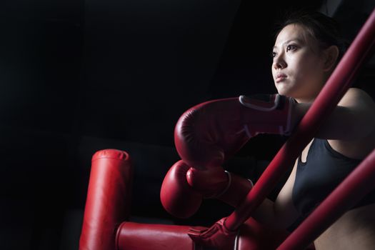 Serious female boxer resting her elbows on the ring side, looking away, low angle view