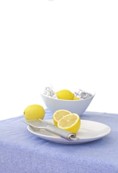 cut lemons on a dish with a lemon zester, with a bowl of lemons in the background. The tablecloth is a light blue, to compliment the yellow of the lemons.