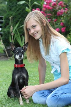 Smiling attractive young teenage girl sitting on green grass with her small dog, a miniature pinscher