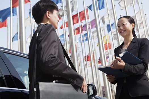 Two businesspeople meeting outdoors with flagpoles in background.