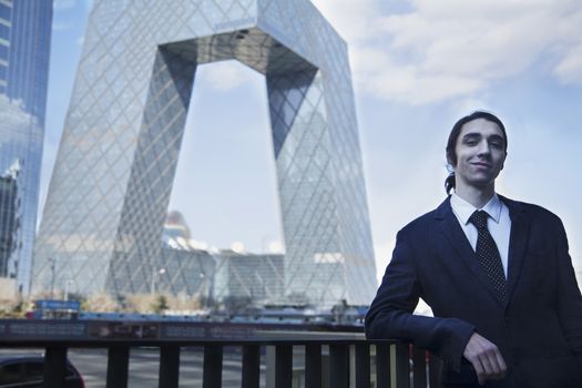 Portrait of smiling young businessman leaning on the railing with the CCTV building in background, Beijing