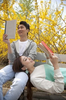 Young happy couple lying and sitting on a park bench enjoying reading their books, outdoors in springtime