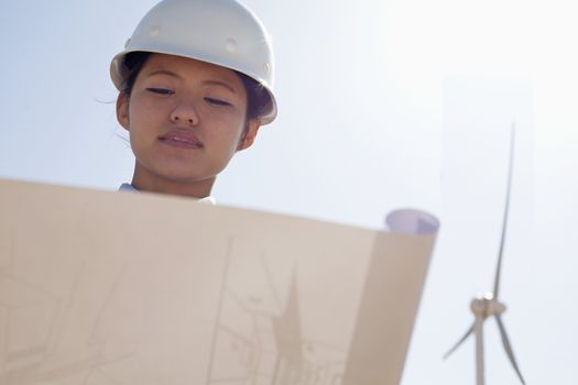 Young female engineer holding open and looking down at blueprints, on site with wind turbines