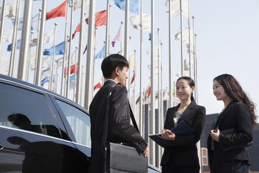 Three businesspeople meeting outdoors with flagpoles in background.