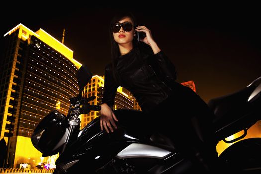 Beautiful young woman with sunglasses talking on the phone and leaning on her motorcycle at night