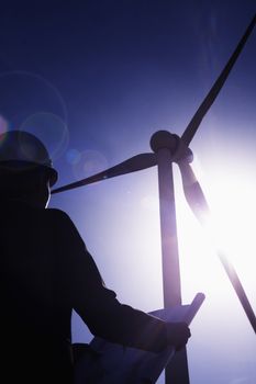 Young female engineer holding open blueprints and checking wind turbines on site, silhouette, low angle view