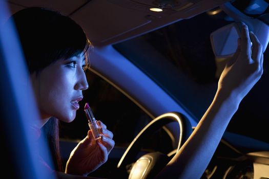 Young beautiful woman in traditional Chinese dress putting on lipstick in the rear view mirror of the car at night