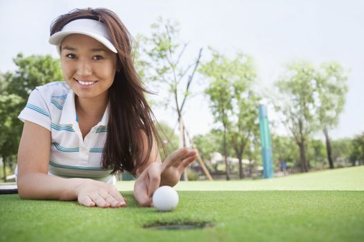 Smiling young woman lying down in a golf course getting ready to flick the ball into the hole