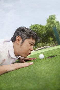 Mid adult man lying down in a golf course trying to blow the ball into the hole