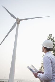 Young male engineer in a hardhat looking up at a wind turbine, blueprint in hand