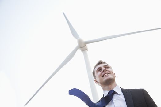 Young smiling businessman standing beside a wind turbine, tie is blowing in the wind