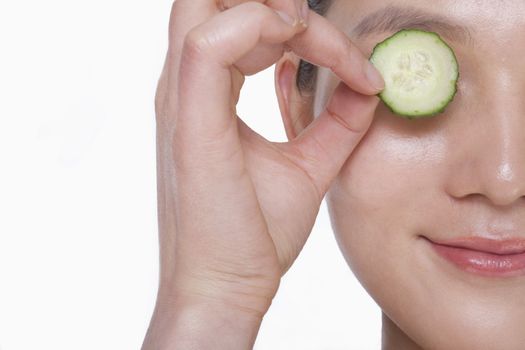 Close up of smiling young woman holding up a cucumber slice over her eye, studio shot