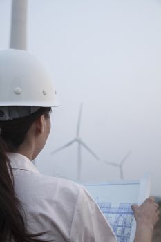 Young female engineer looking at blueprint with wind turbines in front of her, over the shoulder view