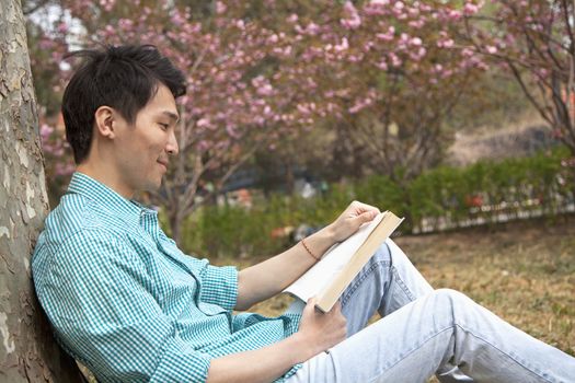 Smiling young man leaning on a tree and enjoying his book, outdoors in a park