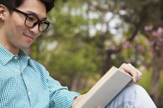 Young man with glasses smiling and enjoying his book, outdoors in a park