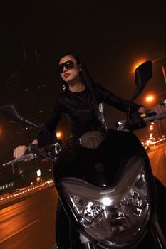 Beautiful young woman riding motorcycle in sunglasses through the city streets at night