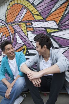 Two young men sitting on their skateboards and hanging out in front of a wall with graffiti