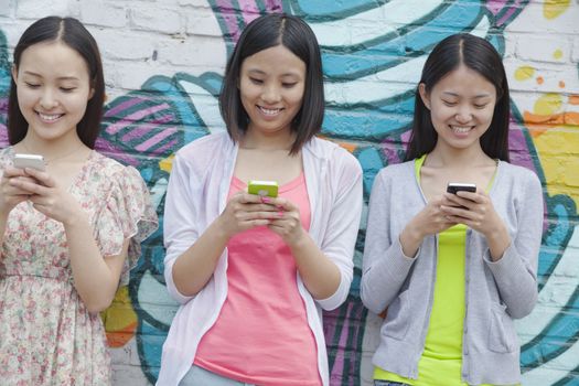 Three smiling young woman standing side by side and texting on their phones in front of a wall with graffiti